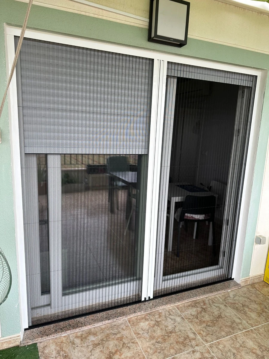 Puerta corredera blanca con persiana y mosquitera plisada en el porche de una vivienda