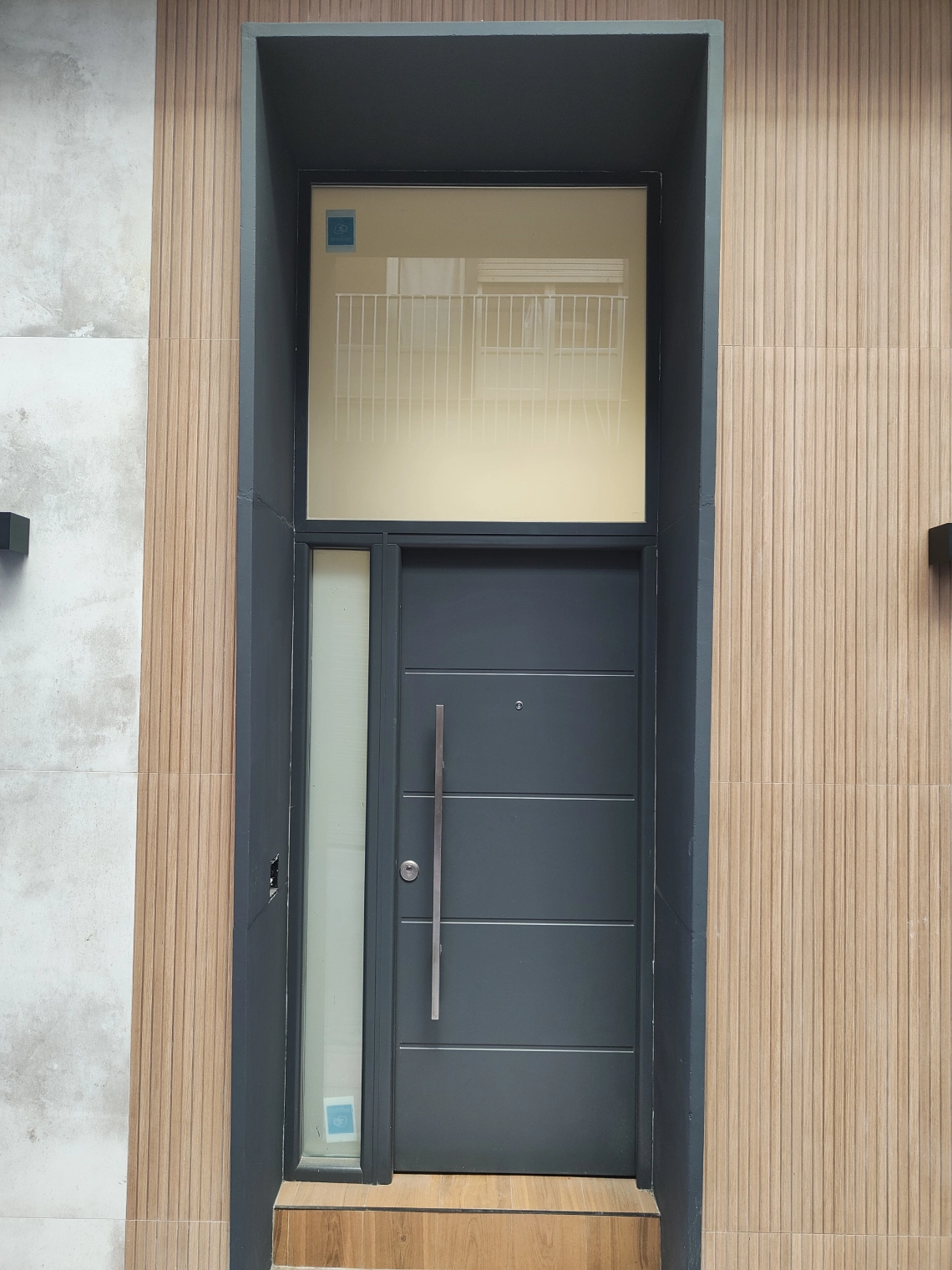 Puerta de entrada de aluminio gris antracita con fijo superior junto a revestimiento de madera
